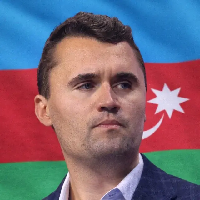 A man in a suit and tie stands in front of a flag.